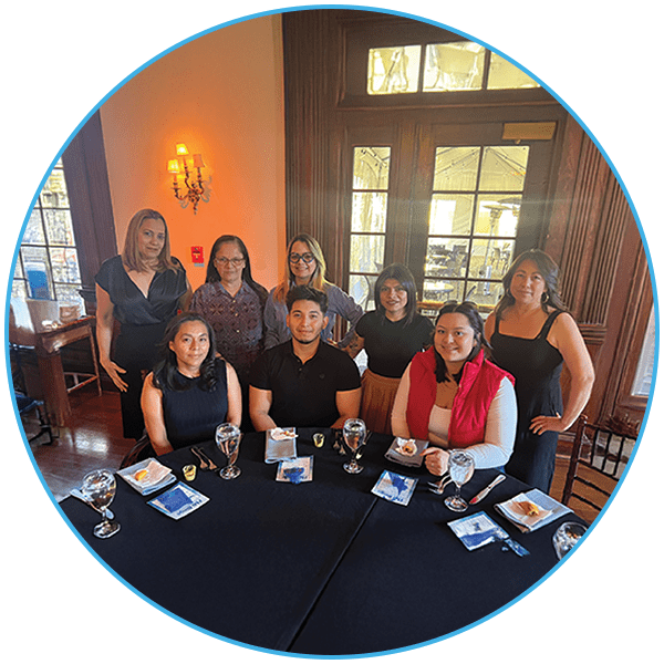 A group of nine people in a formal dining room, seated and standing around a table with plates and glasses.
