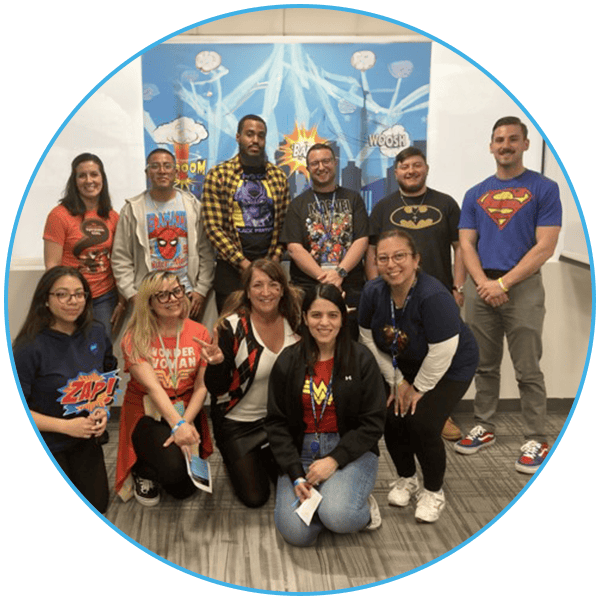 A group of eleven people posing in front of a comic book-themed backdrop, wearing superhero logo T-shirts.