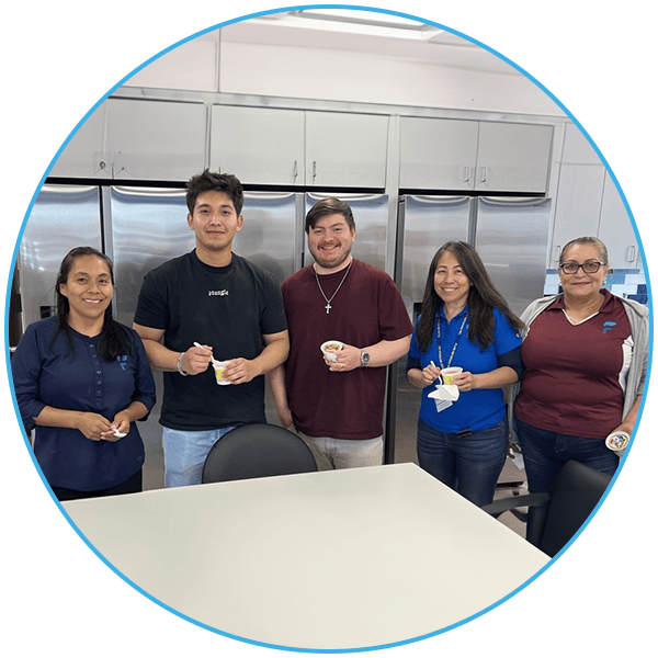 Five people stand in a break room holding ice cream cups in front of a row of stainless steel refrigerators.
