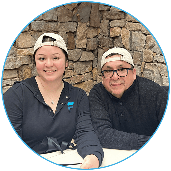 Two individuals wearing backward baseball caps seated in front of a stone wall, both smiling.