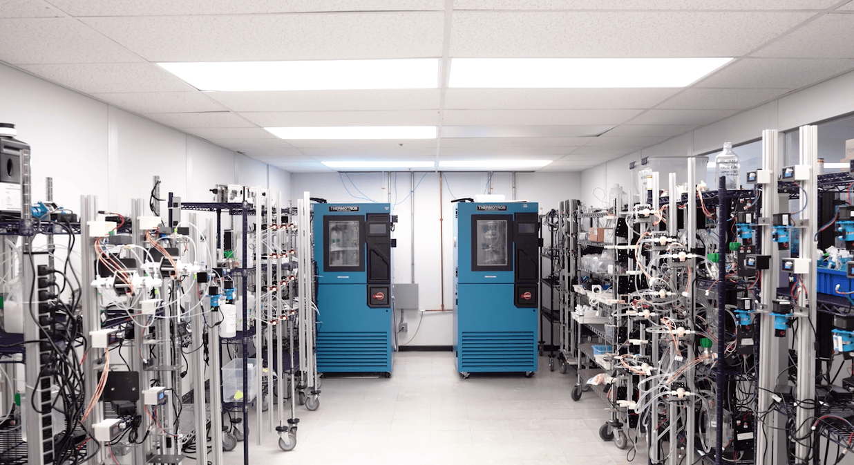 A laboratory room with two blue THERMOTRON machines in the center, surrounded by metal racks filled with pumps and tubes on both sides