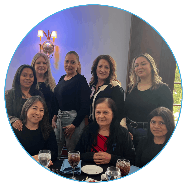 A group of eight women, smiling, pose together indoors near a table.