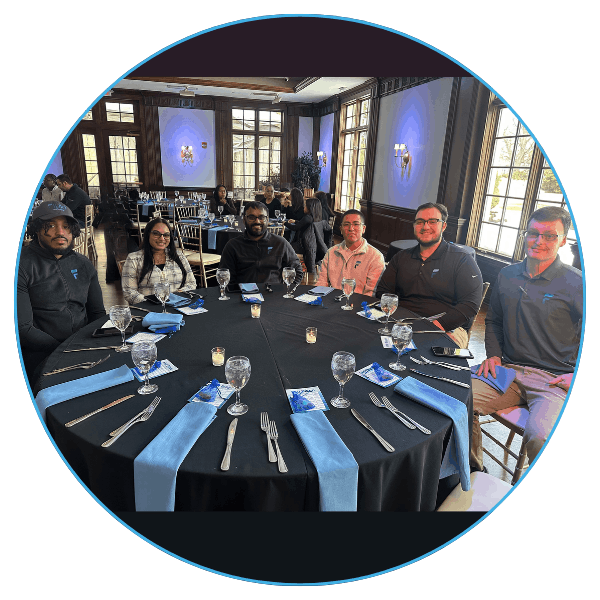 Six people sitting around a round table with a black tablecloth, neatly set with blue napkins, glassware, and silverware, in a well-lit banquet room.