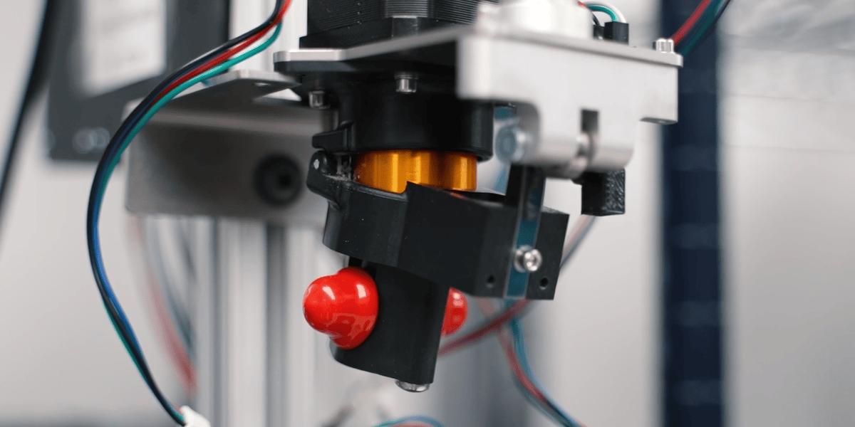 Close-up of a mechanical component with a black plastic part and a red knob, attached to an assembly with an orange cylindrical section and colorful wires