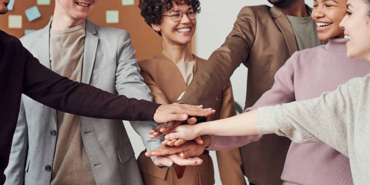 A group of six people smiling and stacking their hands together in a gesture of teamwork